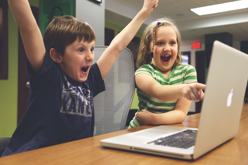 Två barn sitter framför en bärbar dator. Båda ser jätteglada ut. Den ena ler stort och pekar på skärmen. Den andra håller armarna över huvudet och ropar glatt.
