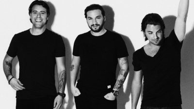 Swedish House Mafia står framför en vit vägg. De står bredvid varandra och har svarta t-shirts på sig. En skrattar, en ler lite försiktigt och en tittar bort och sträcker upp sin arm i luften.